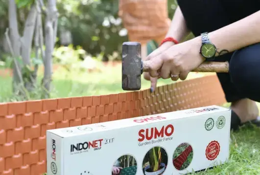 person installing the Indonet Sumo garden border fence into the ground using a mallet, with the product packaging visible in the foreground.